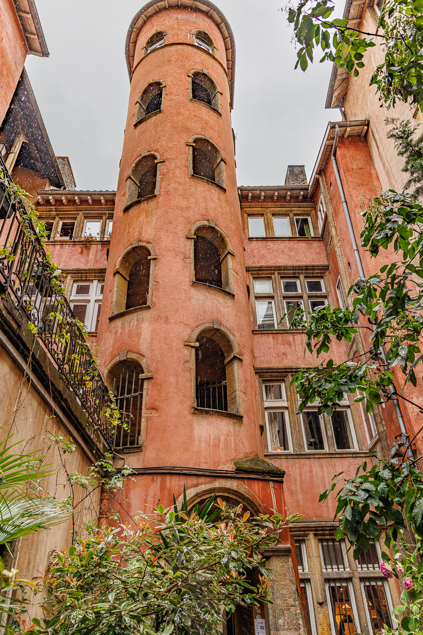 Das Bild zeigt den berühmten rosa Treppenturm des Maison du Crible in der Altstadt von Lyon. Dieses historische Gebäude aus dem 16. Jahrhundert befindet sich in der Rue du Bœuf und ist für seine markante ockerfarbene Fassade bekannt. Der Turm ist ein zentrales Element einer der bekannten Traboules (versteckte Durchgänge) im Viertel Vieux Lyon.