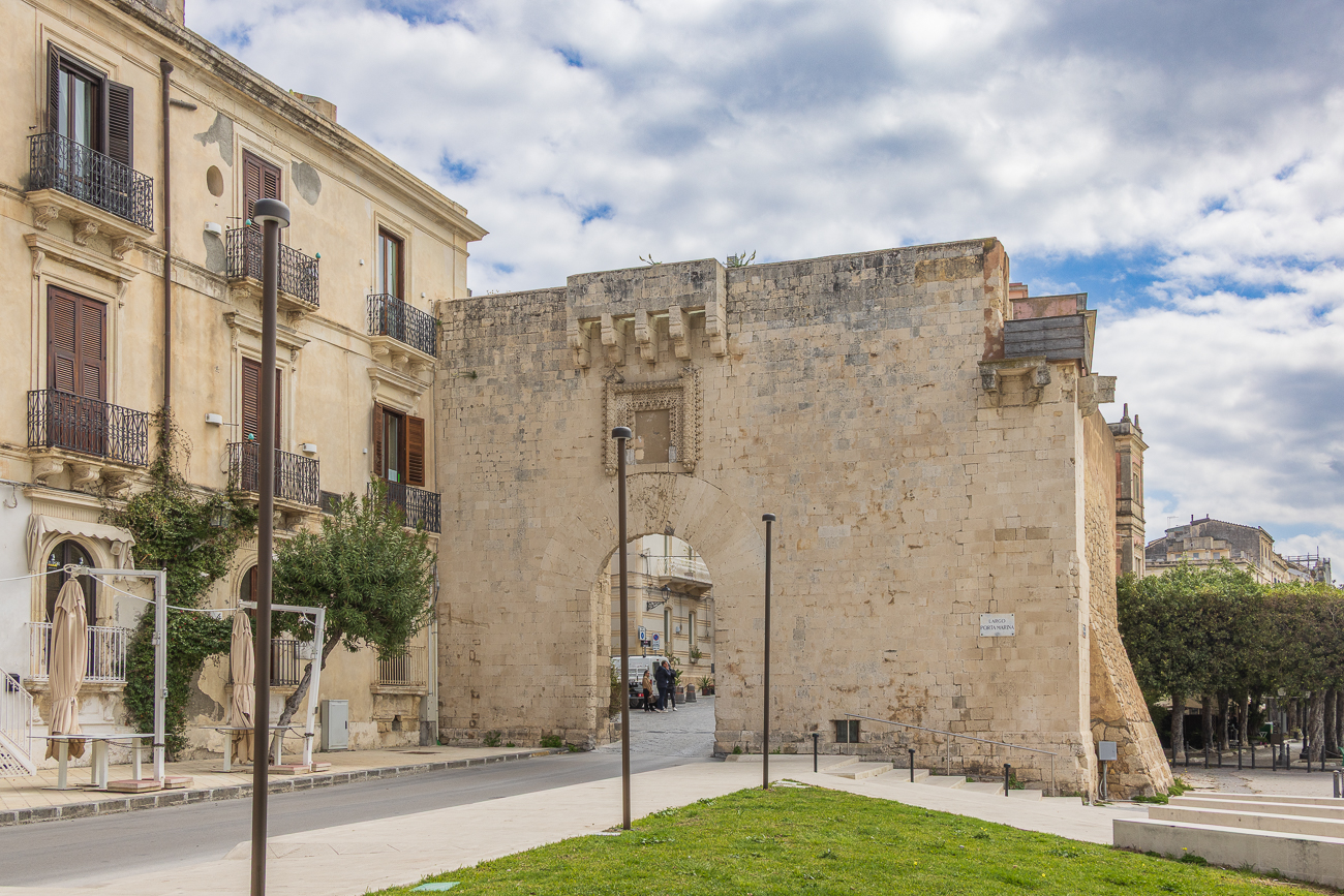 Ein prächtiger Eingang zur Stadt im spanischen Stil, elegant und mächtig zugleich. In den engen Gassen des mittelalterlichen Viertels der Insel Ortygia gelegen, bewahrt das Tor Marina, das den Zugang zur Stadt durch die alten spanischen Befestigungsanlagen ermöglichte, Spuren der katalanischen Epoche in dem der Stein in jedem bearbeiteten und durchbrochenen Teil zu einem floralen Element wird.